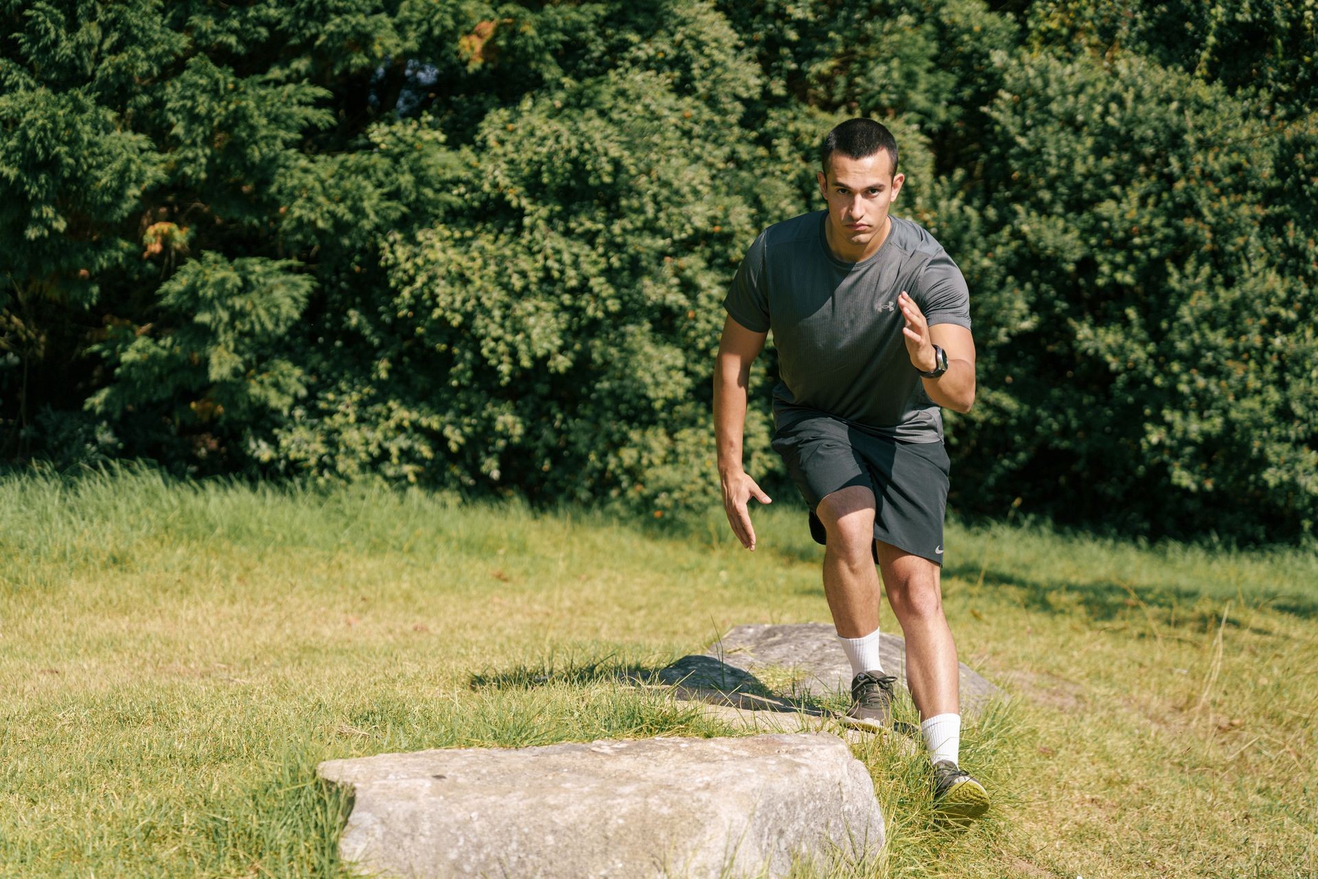 Man exercising outdoors, stepping over large rocks on a grassy field with trees in the background.