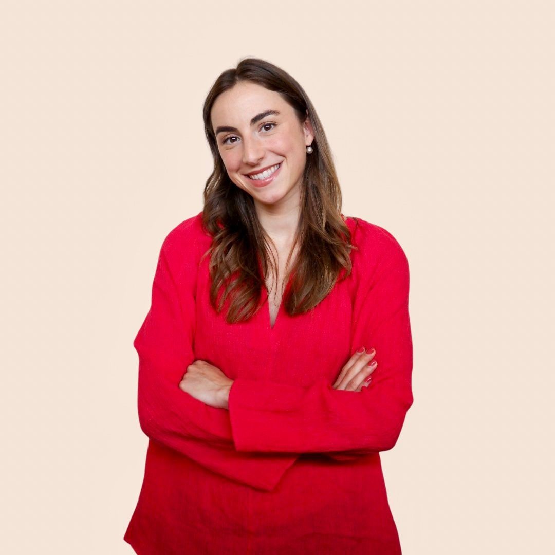 Smiling woman in a red blouse standing with arms crossed against a light beige background.